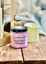 Load image into Gallery viewer, A jar of Mammy's Soap Co. 'Black Velvet' foaming body scrub sits on a rustic wooden table next to a lit mint green candle, with a softly blurred background.