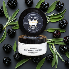 Load image into Gallery viewer, A jar of Mammy's Soap Co. Whipped Blackberry Harvest Body Butter is shown with its lid propped open, surrounded by fresh blackberries and sage leaves on a dark gray background.