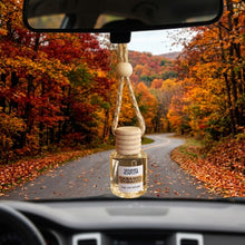 Load image into Gallery viewer, A small glass car air freshener bottle labeled 'Mammy's Soap Co Caramel Tobacco' hangs from a rope attached to a rearview mirror inside a car, with a forest road in autumn visible through the windshield.