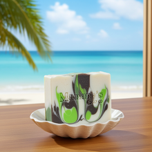 Load image into Gallery viewer, A rectangular bar of soap with a white base and green and dark brown swirls sits on a scalloped beige dish on a wooden table, with a blurred beach and palm tree background.
