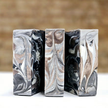Load image into Gallery viewer, Three bars of black, gray, white and brown soap bars with a brown background