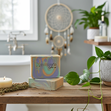 Load image into Gallery viewer, Two decorative handmade soap bars stacked on a rustic wooden table in a bathroom setting. The top bar is marbled with lavender, blue, and gold swirls and has the text 'Mammy's Soap Co' printed on it.