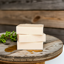 Load image into Gallery viewer, Three stacked rectangular bars of pale cream soap on a rustic wooden board, with a small green sprig and a drop of honey nearby against a blurred wooden plank background.