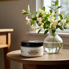 Load image into Gallery viewer, A jar of whipped honeysuckle body butter sits on a wooden table next to a glass vase filled with white flowers and green leaves, in a softly lit indoor setting.