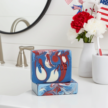 Load image into Gallery viewer, A rectangular bar of soap with a blue, red, and white marbled swirl pattern on a white bathroom counter. The soap has the brand name 'Mammy's Soap Co.
