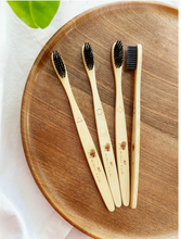 Load image into Gallery viewer, A set of bamboo charcoal toothbrushes with medium/soft bristles, displayed on a wooden plate. The handles of the toothbrushes are etched with different shapes for identification.