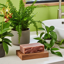 Load image into Gallery viewer, A rectangular pink soap bar resting on a wooden soap dish on a bathroom counter, surrounded by potted green plants and a white sink in a bright bathroom setting.