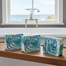 Load image into Gallery viewer, Three handmade soap bars with a blue, green, and white marbled swirl pattern, printed with 'Mammy's Soap Co.' displayed on a wooden ledge in front of a bathtub with a window view of the ocean.