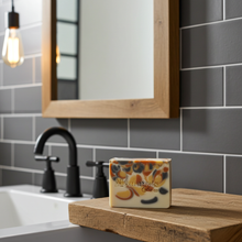 Load image into Gallery viewer, A rectangular bar of soap with a cream base and colorful swirls sits on a wooden cutting board in a bathroom setting with a sink, faucet, mirror, and gray subway tiles.