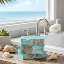 Load image into Gallery viewer, Three stacked turquoise soap bars with coppery rust speckles sit on a wooden surface in front of a sink and window with an ocean view. Seashells and a small potted succulent decorate the scene.