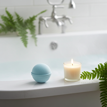 Load image into Gallery viewer, Bath bomb and candle on a bathtub ledge with plants and faucet in the background