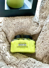 Load image into Gallery viewer, A jar of Sweet Fig sugar body polish with bright yellow-green polish visible through the clear container, resting on a beige crocheted blanket. A black box is partially visible at the top.