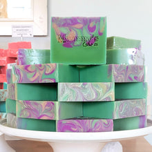 Load image into Gallery viewer, Pedestal plate of stacked green and purple handmade soap bars stamped Mammy&#39;s Soap Co with pink soap bars in the background
