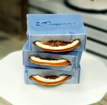 Load image into Gallery viewer, Stack of blue bar soaps with dried orange slices embedded in the top, placed on a white plate with a grey background.