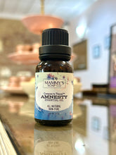 Load image into Gallery viewer, A bottle of Mammy's Soap Co. 'Amnesty' essential oil oil with a black cap and cream label, set on a reflective wooden counter in a softly blurred shop interior.
