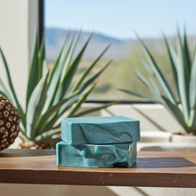 Load image into Gallery viewer, Two teal handmade soap bars stacked on a wooden table, with a blurred desert landscape and agave plants in the background.
