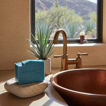 Load image into Gallery viewer, A turquoise bar of Mammy's Soap Co resting on a stone dish on a bathroom counter, next to a copper vessel sink and a small potted succulent, with a window showing a desert landscape in the background.
