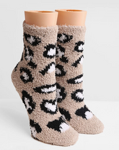 Load image into Gallery viewer, A pair of plush, fuzzy crew socks with a black and white leopard print pattern on a beige background, displayed on peach-colored mannequin legs against a light gray studio background.

