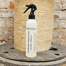 Load image into Gallery viewer, White bottle with black spray nozzle labeled 'Luxury Body Oil Caramel Tobacco' on a wooden surface with a stone wall background.
