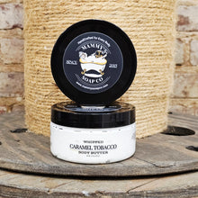 Load image into Gallery viewer, Body butter container with black lid and white body on a wooden surface with jute background
