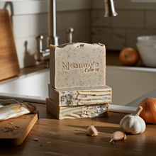 Load image into Gallery viewer, Three stacked handmade bars of soap on a wooden kitchen counter, with a sink, faucet, garlic, onions, and fish in a warm, rustic kitchen setting. The top bar is embossed with the brand name 'Mammy's Soap Co'.
