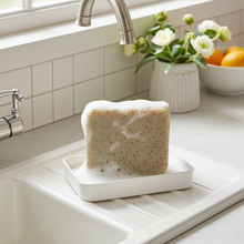 Load image into Gallery viewer, A rectangular white soap dish holding a thick, speckled beige soap bar covered in foam, set in a bright kitchen sink with a tiled backsplash, faucet, flowers, and fruit in the background.
