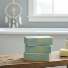 Load image into Gallery viewer, Three stacked rectangular bars of handmade soap in a light green and blue marbled pattern, resting on a wooden bath mat in a bathroom setting with a bathtub and dreamcatcher blurred in the background.
