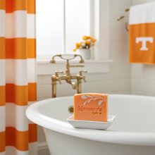 Load image into Gallery viewer, An orange bar of soap labeled 'Mammy's Soap Co' sits on a small white dish on the edge of a white bathtub in a bright bathroom. A matching orange and white striped shower curtain hangs on the left, and an orange towel with a white letter T is visible in the background.
