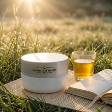 Load image into Gallery viewer, A white cosmetic jar labeled 'Mammy's Soap Co African Rose' sits on a woven mat in grass outdoors, with a glass of golden liquid and an open book nearby.
