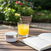 Load image into Gallery viewer, A small white cosmetic jar labeled 'AFRICAN ROSE' sits on a round wooden table outdoors, next to a glass of orange juice and an open book, with a blurred garden background.

