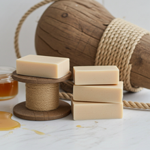 Load image into Gallery viewer, Three rectangular bars of creamy beige soap arranged on a white marble surface, with two spools of twine holding one bar each and a small jar of amber honey in the background. A large wrapped wooden object and rope are also visible behind the setup.

