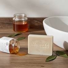Load image into Gallery viewer, A bar of handmade soap from Mammy's Soap Co sits on a wooden surface next to a spilled jar of honey, with a white ceramic bowl in the background.
