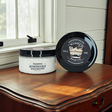 Load image into Gallery viewer, A jar of whipped honeysuckle body butter and its lid placed beside it on a wooden dresser near a window. The products are branded Mammy's Soap Co.
