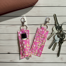 Load image into Gallery viewer, Two pink floral lip balm holders with silver keyrings and clips, shown on a white wood plank surface with keys and a brown leather wallet nearby.
