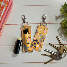 Load image into Gallery viewer, Two fabric lip balm cases with metal clasp closures, displayed on a white wood background alongside keys, a lip balm tube, a pink zip pouch, and a small plant.
