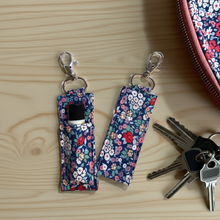 Load image into Gallery viewer, Two fabric lip balm holders with a navy blue floral print and silver lobster clasp rings, shown on a light wood tabletop with keys and part of a matching floral zippered case nearby.
