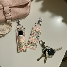 Load image into Gallery viewer, Two fabric lip balm pouches with a pink and beige bohemian pattern, shown with keys and a phone on a light-colored tabletop next to a pink bag.
