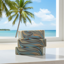 Load image into Gallery viewer, Three stacked rectangular bars of soap with a blue, beige, and brown marbled pattern, displayed on a white windowsill with a tropical beach and palm tree background.

