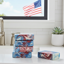 Load image into Gallery viewer, Four rectangular bars of soap with a red, white, and blue marbled pattern are arranged on a white countertop in a bright bathroom setting. A stack of three bars stands on the counter, one bar sits beside them, and a fifth bar is placed in the foreground.
