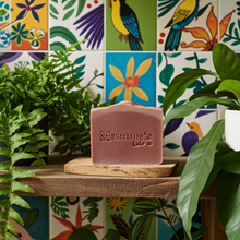 Load image into Gallery viewer, A rectangular bar of dusty rose soap from 'Mammy's Soap Co' sits on a small wooden slice on a rustic wooden shelf, surrounded by green houseplants in front of a colorful tiled wall.
