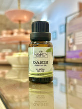 Load image into Gallery viewer, A small amber glass bottle of Mammy's Soap Co. 'Oasis' essential oil with a black cap and a white label, set on a reflective glass counter with a blurred shop background.
