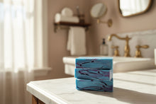 Load image into Gallery viewer, A bar of artisan soap with a blue and purple swirl design, placed on a tiled surface with soft lighting in the background.
