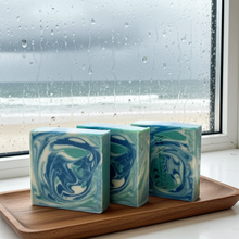 Load image into Gallery viewer, Three rectangular handmade soap bars with blue, green, and white marbled swirls, displayed on a wooden tray in front of a rain-streaked window overlooking the beach.
