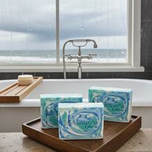 Load image into Gallery viewer, Three bars of Mammy's Soap with blue and white marbled swirls, displayed on a wooden tray in front of a bathtub with a window showing rain and the ocean outside.
