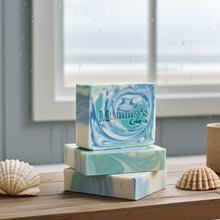 Load image into Gallery viewer, Three stacked rectangular bars of soap with blue and white marbled swirls, featuring the brand name 'Mammy's Soap Co' on the top bar. The soap bars are arranged on a wooden surface next to seashells, with a window and raindrops in the background.
