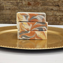 Load image into Gallery viewer, Three bars of bronze, gold and white soaps with swirls on a gold plate with a brick wall in the background.
