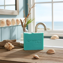 Load image into Gallery viewer, A turquoise bar of soap labeled 'Mammy's Soap Co.' sits on a rustic wooden surface outdoors near a window with a view of the ocean, styled with seashells and driftwood.
