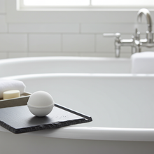 Load image into Gallery viewer, White bath bomb on a black soap dish in a bathtub with a tiled wall and faucet in the background.
