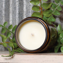 Load image into Gallery viewer, Candle in a brown jar surrounded by green leaves on a wooden surface
