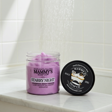 Load image into Gallery viewer, A jar of Mammy's Soap Co. 'Starry Night' foaming body scrub with purple scrub visible through a clear jar, shown next to its black lid on a white marble surface with water droplets falling in the background.
